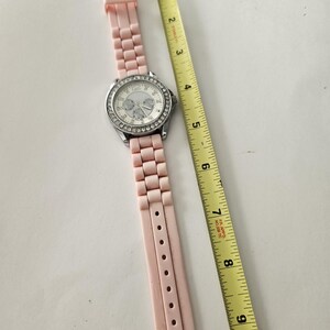 Op de afbeelding: Een roze polshorloge met een zilverkleurige lunette en een lichtroze band. De wijzerplaat is wit met zilverkleurige wijzers en markeringen. Een gele meetlint ligt naast het horloge. De band is ongeveer 20 cm lang.