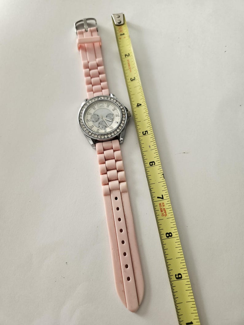 Op de afbeelding: Een roze polshorloge met een zilverkleurige lunette en een lichtroze band. De wijzerplaat is wit met zilverkleurige wijzers en markeringen. Een gele meetlint ligt naast het horloge. De band is ongeveer 20 cm lang.