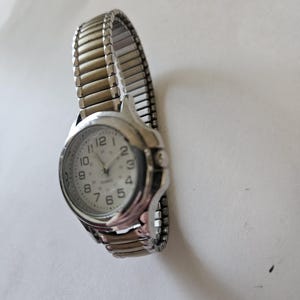 May include: A silver-toned quartz watch with a white face and black numerals. The watch has a stretch band and a round face. The watch hands are black. The watch is a classic style.
