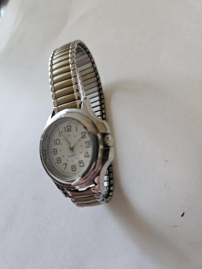 May include: A silver-toned quartz watch with a white face and black numerals. The watch has a stretch band and a round face. The watch hands are black. The watch is a classic style.