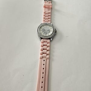 Op de afbeelding: Een polshorloge met een lichtroze band en een zilverkleurige wijzerplaat. De wijzerplaat is omgeven door een ring van kleine, heldere stenen. De horlogeband is gemaakt van een flexibel materiaal.