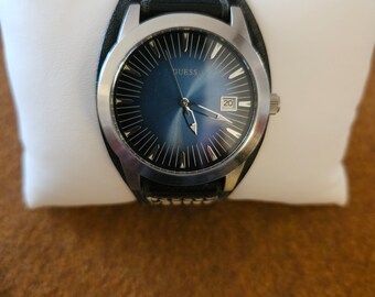 Puede incluir: Un reloj Guess plateado con una esfera azul y una correa de cuero negro. El reloj tiene una ventana de fecha en la posición de las 4 en punto.