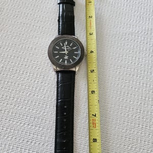 May include: A black wristwatch with a silver-colored bezel and a black leather band. The watch face is black with silver-colored hands and markers. The watch is approximately 9 inches long.