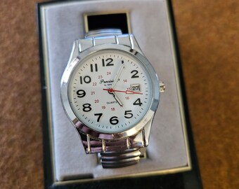 Puede incluir: Un reloj de pulsera plateado con una esfera blanca y números negros. El reloj tiene una correa de metal plateado y una ventana de fecha en la posición de las 3 en punto. La esfera del reloj dice "Precisi by GRE" y "QUARTZ".