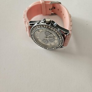 Op de afbeelding: Een zilverkleurig polshorloge met een lichtroze siliconen band. De wijzerplaat van het horloge is wit met drie sub-wijzerplaten en een bezel bezet met kleine, heldere kristallen. De band is vastgezet met een zilveren gesp.