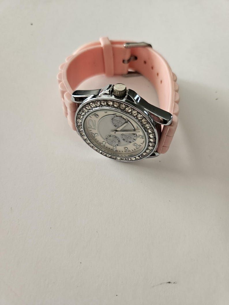Op de afbeelding: Een zilverkleurig polshorloge met een lichtroze siliconen band. De wijzerplaat van het horloge is wit met drie sub-wijzerplaten en een bezel bezet met kleine, heldere kristallen. De band is vastgezet met een zilveren gesp.