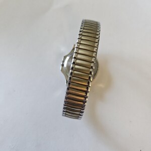 May include: A silver-toned wristwatch with a gold-toned, expandable band. The watch face is round and the band is made of small, rectangular metal links. The watch is set against a white background.