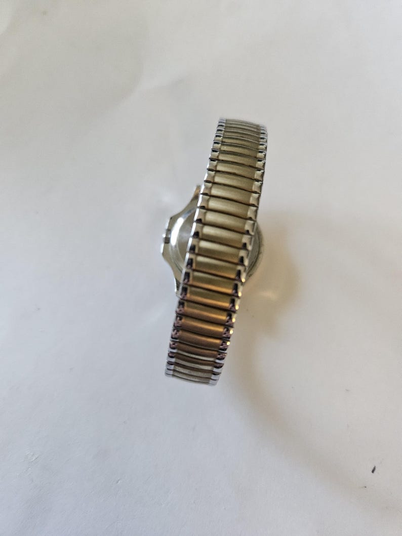 May include: A silver-toned wristwatch with a gold-toned, expandable band. The watch face is round and the band is made of small, rectangular metal links. The watch is set against a white background.