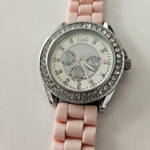 Op de afbeelding: Een zilverkleurig horloge met een witte wijzerplaat en een roze band. De wijzerplaat heeft drie sub-wijzerplaten en is omgeven door een ring van heldere kristallen. De roze band is gemaakt van flexibel materiaal.