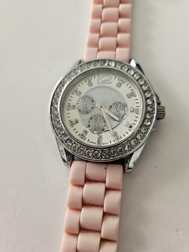 Op de afbeelding: Een zilverkleurig horloge met een witte wijzerplaat en een roze band. De wijzerplaat heeft drie sub-wijzerplaten en is omgeven door een ring van heldere kristallen. De roze band is gemaakt van flexibel materiaal.
