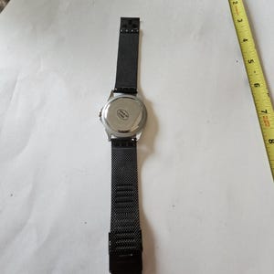 May include: A black wristwatch with a mesh metal strap and a silver-toned circular case back. The watch is positioned next to a yellow measuring tape, showing a length of approximately 8 inches from the top of the strap to the bottom.