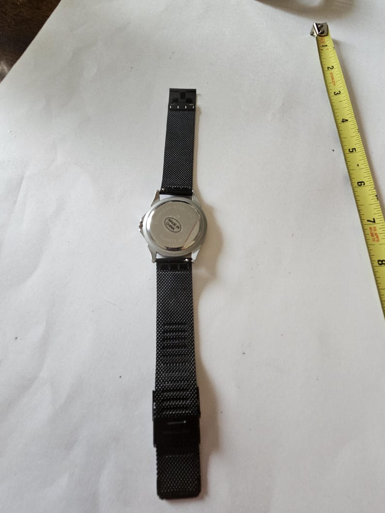 May include: A black wristwatch with a mesh metal strap and a silver-toned circular case back. The watch is positioned next to a yellow measuring tape, showing a length of approximately 8 inches from the top of the strap to the bottom.