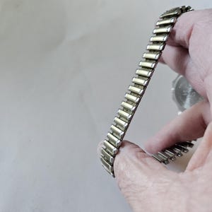 May include: A silver-toned, expandable watch band with rectangular links. The band is held in a hand, showcasing its flexible design. The watch face is partially visible in the background.