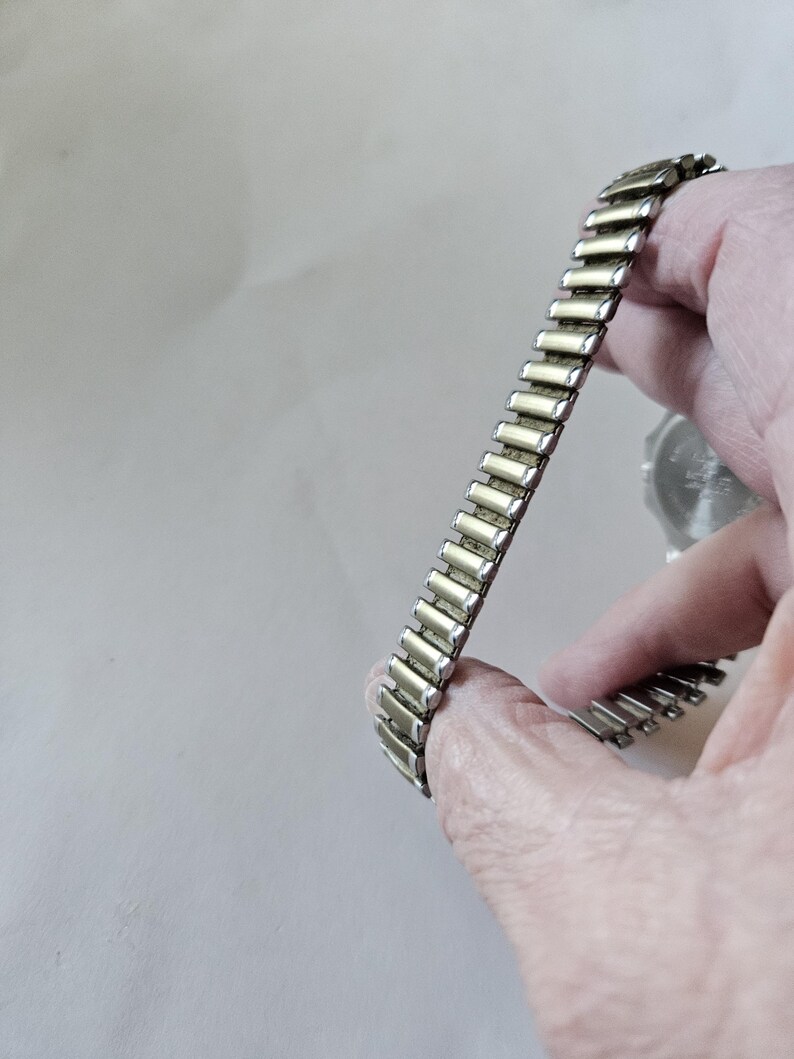 May include: A silver-toned, expandable watch band with rectangular links. The band is held in a hand, showcasing its flexible design. The watch face is partially visible in the background.
