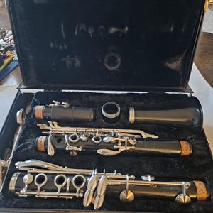 May include: A black clarinet in a black case. The instrument is disassembled, with the various parts neatly arranged within the case. The keys and other hardware are silver-colored. The case is open, revealing the instrument's components.