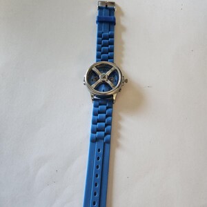 Puede incluir: Un reloj de pulsera azul con caja plateada y correa azul a juego. La esfera del reloj presenta un dise&ntilde;o &uacute;nico con forma de "X" y n&uacute;meros visibles. La correa es de material flexible y tiene varios orificios de ajuste.