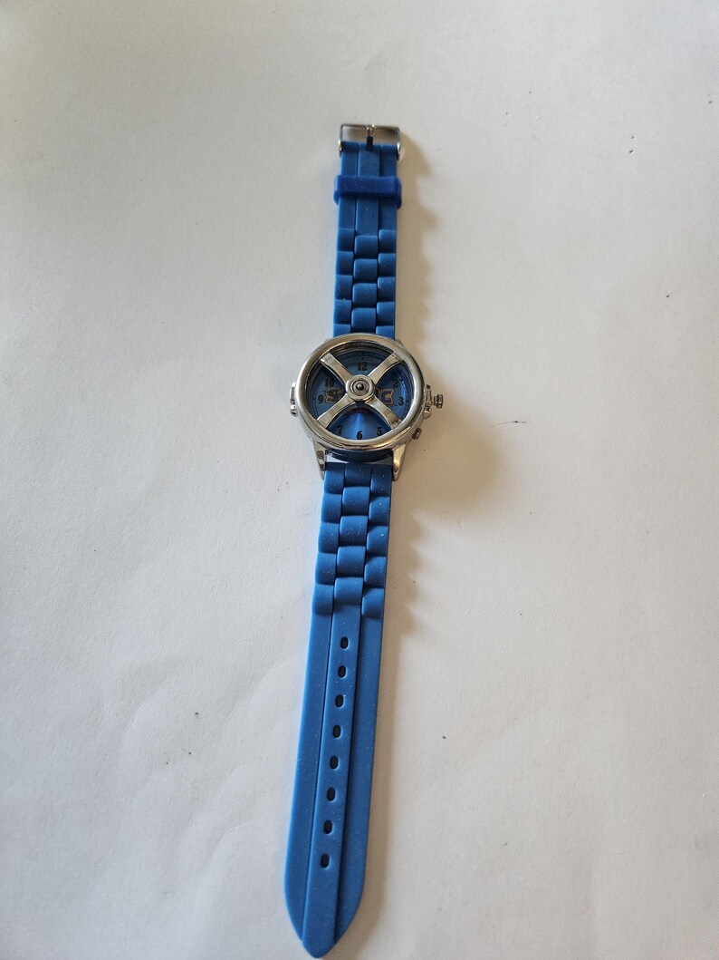 Puede incluir: Un reloj de pulsera azul con caja plateada y correa azul a juego. La esfera del reloj presenta un dise&ntilde;o &uacute;nico con forma de "X" y n&uacute;meros visibles. La correa es de material flexible y tiene varios orificios de ajuste.