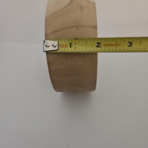 Puede incluir: Una pata de mueble de madera clara, de aproximadamente 5 cm de di&aacute;metro, se mide con una cinta m&eacute;trica amarilla. La pata tiene forma cil&iacute;ndrica con parte superior e inferior planas. Se aprecia la veta de la madera.