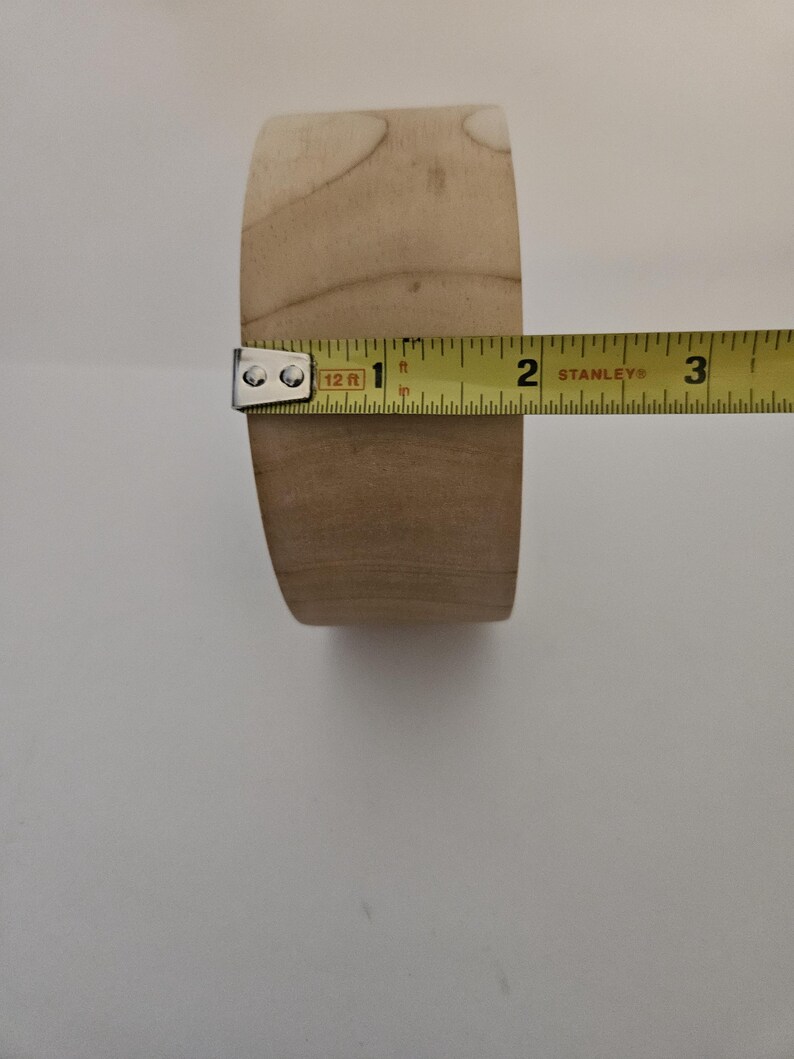 Puede incluir: Una pata de mueble de madera clara, de aproximadamente 5 cm de di&aacute;metro, se mide con una cinta m&eacute;trica amarilla. La pata tiene forma cil&iacute;ndrica con parte superior e inferior planas. Se aprecia la veta de la madera.