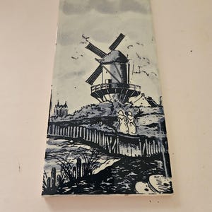 May include: A rectangular ceramic tile featuring a blue and white illustration of a traditional windmill situated on a grassy hill. Two figures wearing bonnets stand near a wooden fence by the water, with a small building visible in the distance.