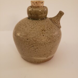 May include: A small, light brown ceramic bottle with a cork stopper. The bottle has a rounded body and a small spout. It has a speckled glaze and a rustic look.