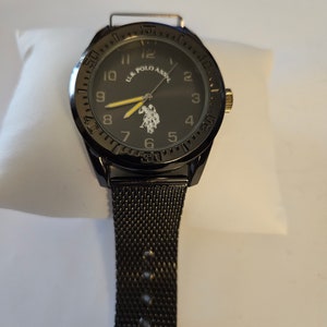 May include: Black wristwatch with a black mesh band. The watch face is black with white numbers and a logo that says "U.S. Polo Assn."