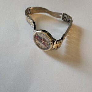 May include: A wristwatch with a silver and gold-tone metal band. The watch face has a colorful design with red, blue, and white accents. The watch is angled, showing the face and part of the band.