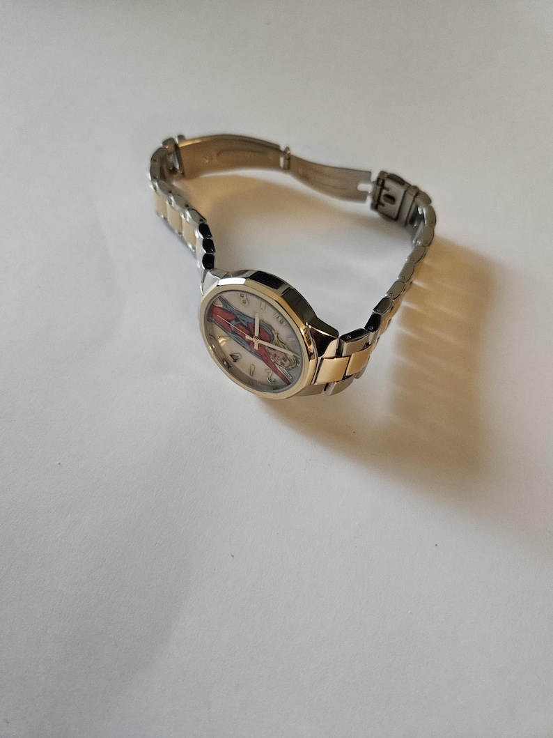 May include: A wristwatch with a silver and gold-tone metal band. The watch face has a colorful design with red, blue, and white accents. The watch is angled, showing the face and part of the band.