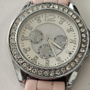 Op de afbeelding: Een zilverkleurig horloge met een witte wijzerplaat, drie sub-wijzerplaten en een bezel bezet met kristallen. Het horloge heeft een lichtroze siliconen band. De wijzerplaat toont het nummer 12 in zilver.