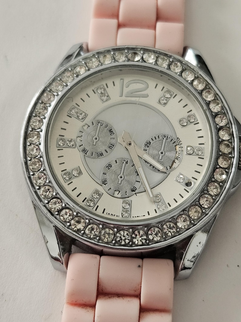 Op de afbeelding: Een zilverkleurig horloge met een witte wijzerplaat, drie sub-wijzerplaten en een bezel bezet met kristallen. Het horloge heeft een lichtroze siliconen band. De wijzerplaat toont het nummer 12 in zilver.