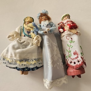 May include: Three small dolls with painted faces and fabric clothing. The left doll wears a white apron over a blue floral skirt. The center doll wears a light blue gown with white floral embroidery. The right doll wears a white dress with red embroidery.