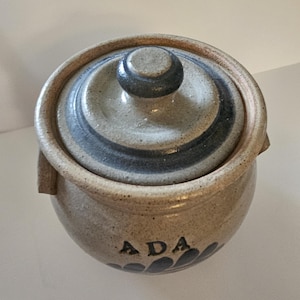 May include: A ceramic jar with a lid, featuring a speckled beige finish and dark blue stripes on the lid. The word "ADA" is printed on the jar's side, with a floral design below. The jar has a round shape and a small handle.