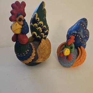 May include: Two decorative rooster figurines. The larger rooster has a red comb, blue and green feathers, and a yellow beak. The smaller rooster is blue, green, and orange. Both have detailed, textured designs and are set against a white background.