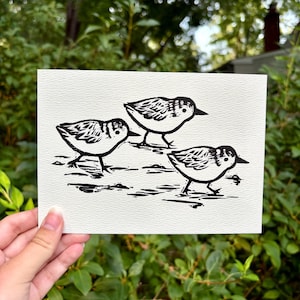 May include: A rectangular white card featuring a black ink block print of three small sandpiper birds. The birds are depicted in a side profile view with textured feather details, standing on a simple ground line against a plain background.