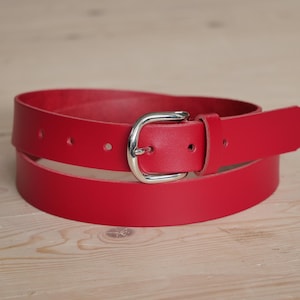 Peut inclure: Une ceinture en cuir rouge avec une boucle argentée. La ceinture est enroulée et la boucle est visible.