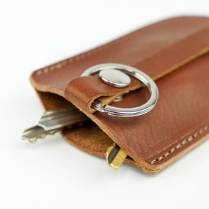 May include: A brown leather key holder with a silver key ring and two keys. The key holder has a stitched edge and a flap that folds over the keys.