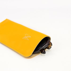 May include: A yellow leather glasses case with a white logo. The case is folded over and a pair of brown sunglasses are partially visible inside.