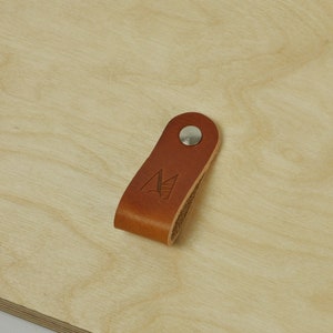 May include: A brown leather strap with a metal button and a logo of a mountain with a sun and waves. The strap is on a light wood surface.