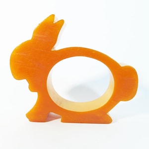 May include: Orange bunny-shaped napkin ring. The ring is made of a solid orange material with a bunny silhouette. The bunny has a long ear, a round body, and a small tail. The napkin ring is designed to hold a rolled-up napkin.