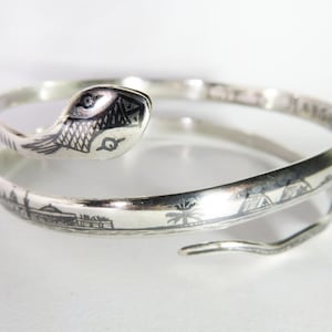 May include: A silver bangle bracelet featuring a snake design. The snake is coiled around the bracelet and has a detailed head with an eye. The bracelet is engraved with a pattern of geometric shapes.