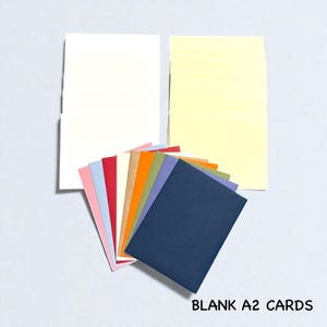 May include: A collection of blank A2 cards in various colors, including white, yellow, pink, red, and blue. The cards are fanned out, with two larger cards in the background. Text at the bottom reads "BLANK A2 CARDS".