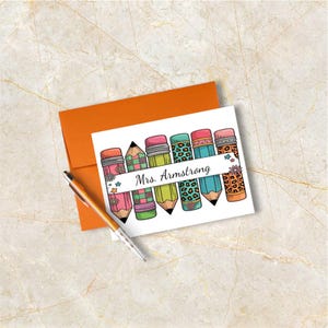 May include: A white greeting card with a row of colorful pencils, each with a unique design, and the text "Mrs. Armstrong". An orange envelope and a pen are also present. The pencils have pink, green, blue, and orange accents.