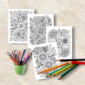 May include: Four white coloring pages with intricate floral designs are displayed. A green cup holds a collection of colored pencils, and several pencils are scattered nearby. The pages feature detailed black and white illustrations of flowers and leaves.