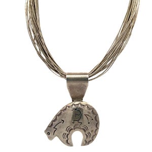 May include: A multi-strand silver liquid necklace featuring a polished silver pendant shaped like a bear. The pendant is etched with a Kokopelli figure and decorative sunburst patterns along the outer edge, suspended from a silver bail.
