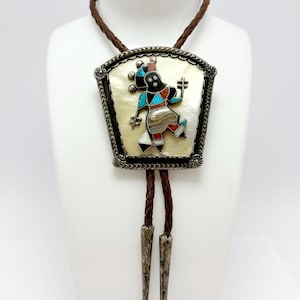 May include: A bolo tie with a rectangular pendant featuring a colorful inlaid figure on a mother-of-pearl background. The pendant is framed with a silver-toned border and hangs from a brown braided leather cord with silver-toned tips.