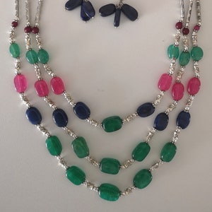 Könnte beinhalten: Eine dreireihige Halskette mit grünen, rosa und blauen ovalen Perlen, silbernen Zwischenstücken und silbernen Endkappen. Passende Ohrringe mit blauen ovalen Perlen und silbernen Akzenten.