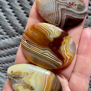 Può includere: Vengono mostrate tre pietre di agata lucidata nei toni del marrone, crema e bianco. Le pietre hanno motivi a bande unici e sono di forma ovale, con un foro praticato nella parte superiore per l'infilatura. Le pietre sono tenute in una mano.