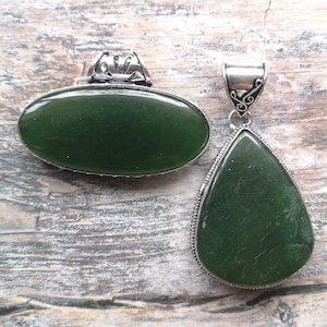 Könnte beinhalten: Zwei Anhänger mit grünen Jade-Edelsteinen. Einer ist oval, der andere tropfenförmig. Beide haben silberfarbene Metalleinfassungen mit dekorativen Details. Der Jade hat eine polierte Oberfläche, das Metall ein antikes Finish.