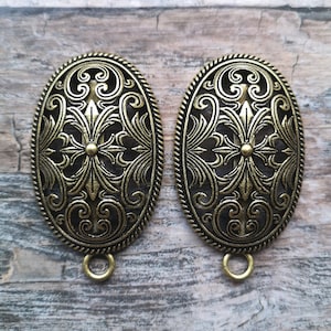 Può includere: Due ornamenti metallici ovali di colore oro antico con intricati disegni floreali. Ogni ornamento ha un piccolo anello nella parte inferiore.