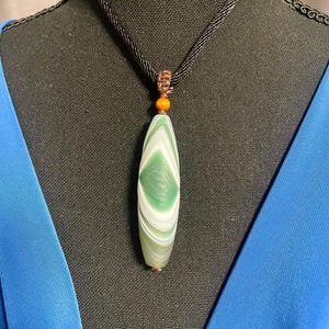 May include: A green and white oval pendant necklace. The pendant is suspended from a black cord and features a marbled design. A small orange bead accents the top of the pendant. The necklace is displayed on a black mannequin.